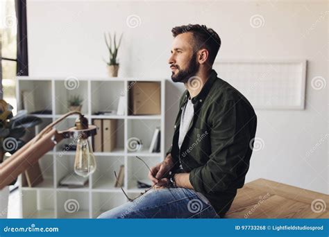 Bearded Man Holding Eyeglasses While Sitting At Table And Looking Away