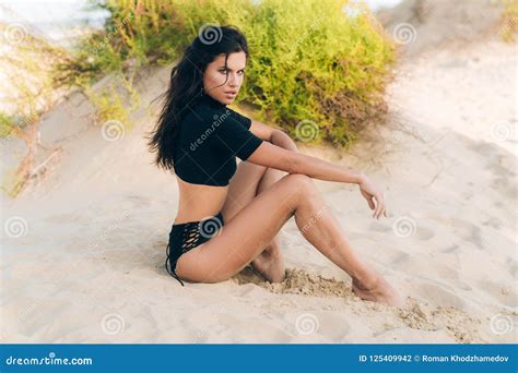 Young Brunette With Curly Hair Curls Fall On The Face Posing Sitting On The Beach Wearing A