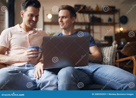 Loving Same Sex Male Couple Sitting On Sofa Using Laptop As They Relax At Home Together Stock