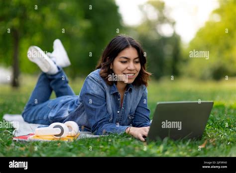Smiling Arab Female Student Using Laptop While Lying On Lawn Outdoors
