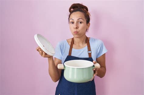 Brunette Woman Wearing Apron Holding Cooking Pot Puffing Cheeks With Funny Face Stock Photo