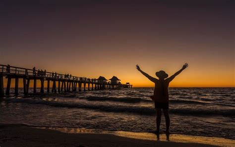 Una Mujer Se Para En La Playa Frente A Un Muelle Con La Puesta De Sol