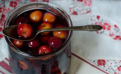 A Jar Filled With Lots Of Plums On Top Of A White Table Next To A Spoon