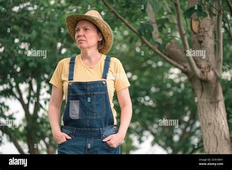 Portrait Of Female Farmer Posing In Walnut Fruit Orchard Wearing Straw Hat And Bib Overalls