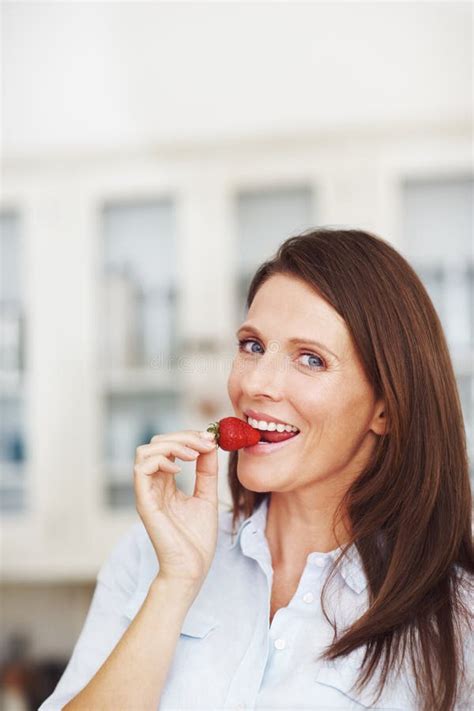 Bite Sized Bliss An Attractive Brunette Eating A Strawberry While Standing In A Kitchen Stock