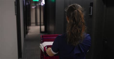 Young Adult Professional Maid Walking And Rolling Trolley In Corridor Of Luxury Hotel Back View