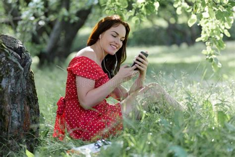 Plus Size Girl Sitting On Grass Under Tree And And Using Her Smartphone