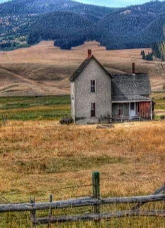 An Old Farm House Sits In The Middle Of A Field With Mountains In The