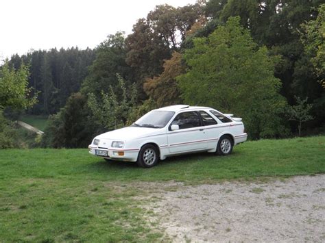 A White Car Parked On Top Of A Lush Green Field Next To Trees And Grass