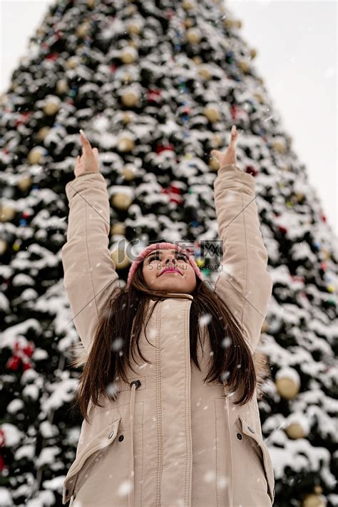 穿着温暖冬衣的女人站在户外的大圣诞树旁双臂高举雪花飘落高清摄影大图 千库网