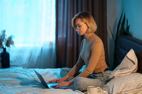 Blonde Frau mit kurzen Haaren auf weißem Bett in Jeans mit einem Laptop