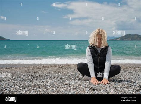 Blonde European Woman Sits On A Pebble Beach On A Spring Day Touches The Stones On The Beach