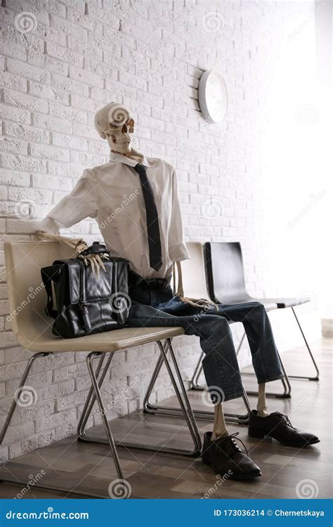 Human Skeleton In Office Wear Sitting On Chair Near Wall Indoors Stock