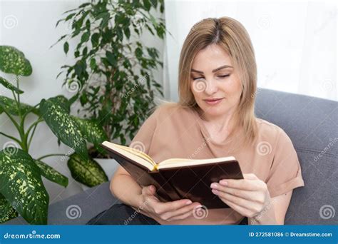 Blonde Woman Reading Book While Sitting On Beige Armchair In Apartment Stock Photo Image Of