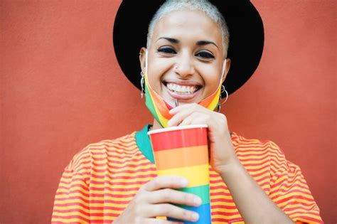 Feliz joven gay bebiendo de un vaso de arco iris mientras usa una máscara de orgullo enfoque
