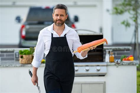 Male Chef In Cook Apron Grilling And Barbequing Salmon Fish In Garden