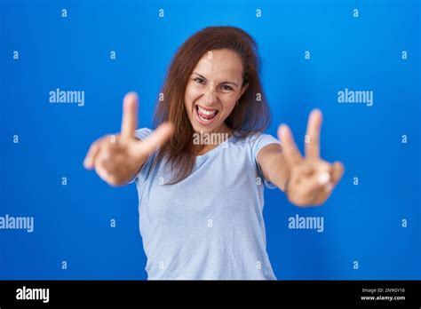 Brunette Woman Standing Over Blue Background Smiling With Tongue Out Showing Fingers Of Both