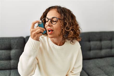 Mature Hispanic Woman Using Asthma Inhaler Sitting On The Sofa At Home
