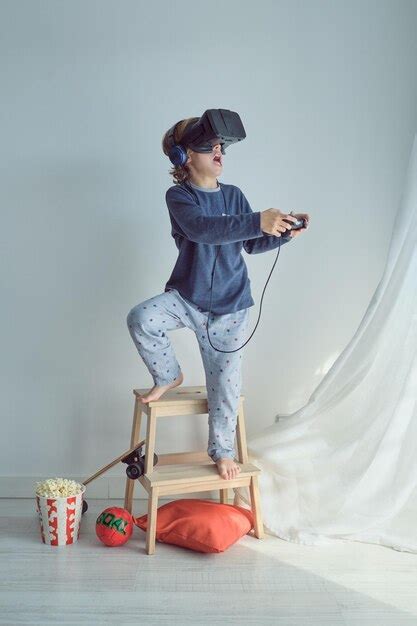 ナイトウェアと vr ヘッドセットとヘッドフォンを着た裸足のプレティーンの少年の全身がスツールの脚立の上に立ってジョイスティックでビデオ