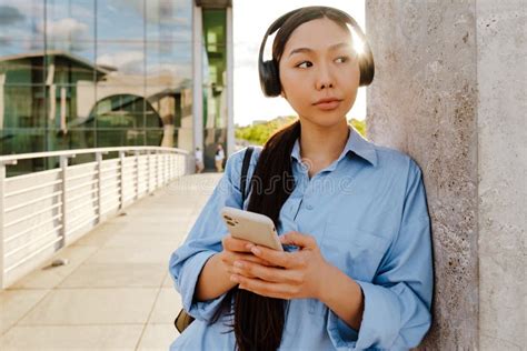 Asian Brunette Woman Using Cellphone While Standing By Wall Stock Photo