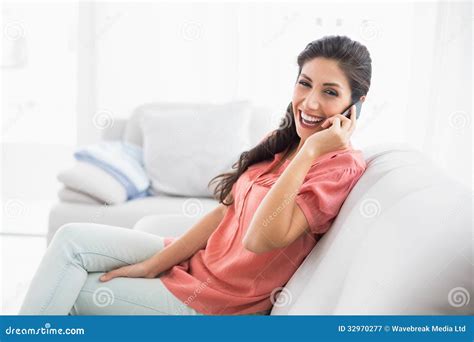 Laughing Brunette Sitting On Her Sofa On The Phone Looking At Ca Stock