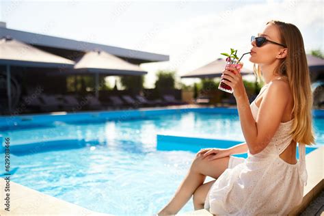 Blonde Woman Drinks Refreshing Cocktail Sunbathing And Sitting Near Swimming Pool At Tropical