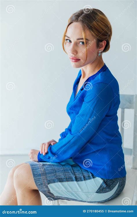 A Woman In Blue Clothes Sits On A Chair Against A White Wall Stock