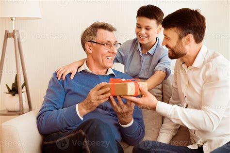 Mature Father And Son Congratulating Grandpa Giving Him Birthday Gift Sitting On Sofa At Home