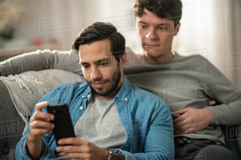 Cute Male Gay Couple Spend Time At Home They Are Lying Down On A Sofa And Use A Smartphone