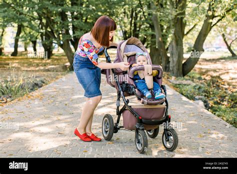 family, child and parenthood concept - happy mother walking with baby stroller in park from back ...