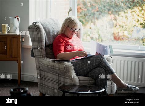 Une femme mature est assise sur un fauteuil dans le salon de sa maison la lecture à travers des