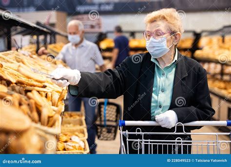 Older Woman With Glasses Chooses Buns And Bread In Supermarket Bakery