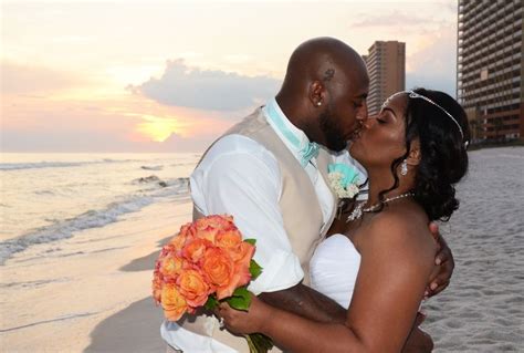 A Bride And Groom Kissing On The Beach At Sunset With Buildings In The