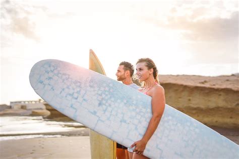 Young Couple Of Happy Surfers Standing On The Beach Holding Surfboards