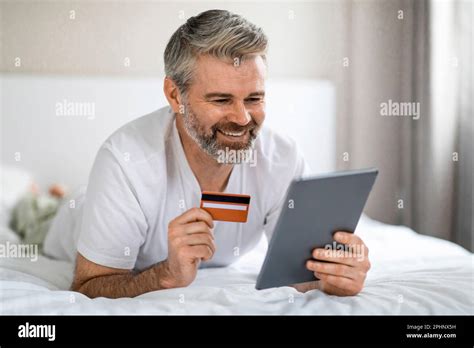 Positive Mature Man Enjoying Online Shopping Lying On Bed Stock Photo