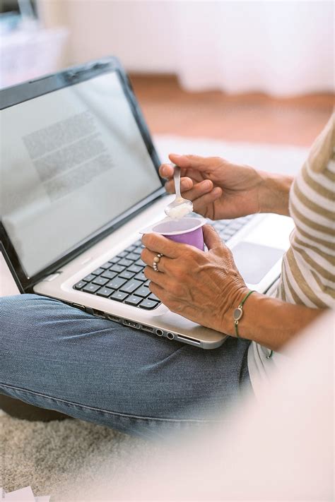 Closeup Of Mature Woman Eats Yogurt While Reading A Document On Laptop By Stocksy Contributor