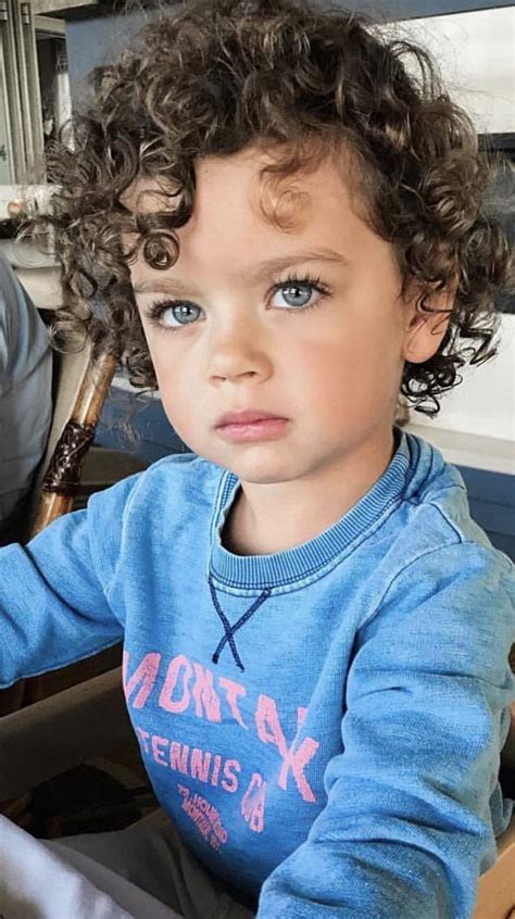 A Little Boy Sitting In A Chair With Curly Hair On His Head And Blue Eyes