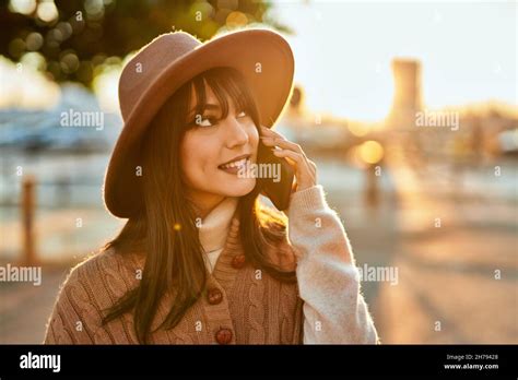 Brunette Woman Wearing Winter Hat Smiling Speaking On The Phone Outdoors At The City On Sunset