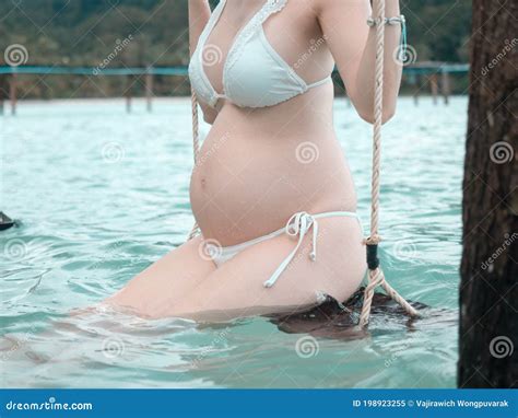 Asian Pregnant Woman In Bikini Sitting On Hammock In Turquoise Sea