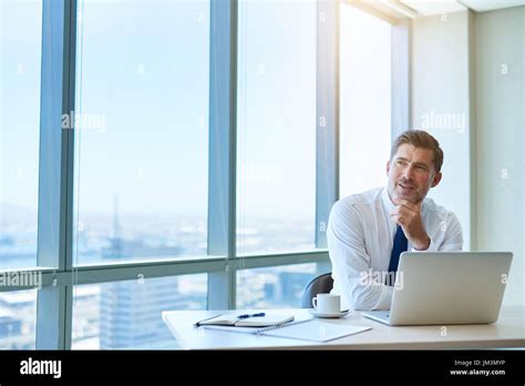 Attractive And Mature Businessman Sitting At His Desk In A Modern Office With Large Windows