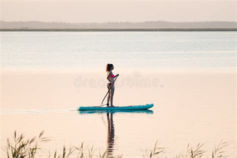 A Woman In Mohawk Shorts Stands On A Sup Board At Sunset In A Lake