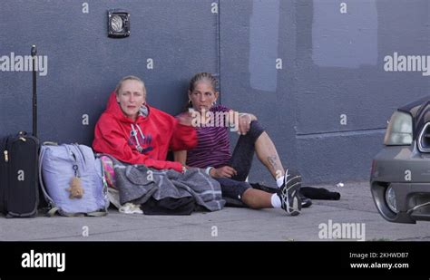 Two Homeless Women Sitting On A Sidewalk Sharing A Cigarette Stock