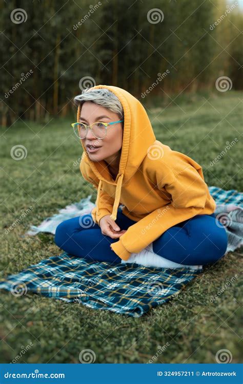 Cute Girl With Glasses In An Orange Hoodie Is Sitting In A Park In