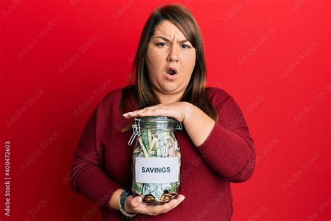 Beautiful Brunette Plus Size Woman Holding Jar With Savings In Shock Face Looking Skeptical And