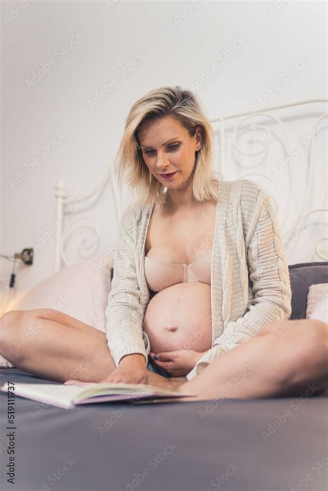 Vertical Indoor Shot Of A Beautiful Blonde Caucasian Pregnant Woman In A Beige Cardigan Touching