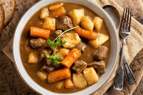 A Bowl Of Stew With Carrots Potatoes And Meat On A Wooden Table Next