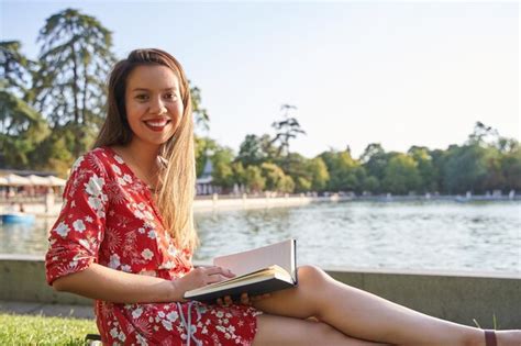 Jovem latina lendo um livro sentado no gramado na margem de um lago olhando para a câmera Foto