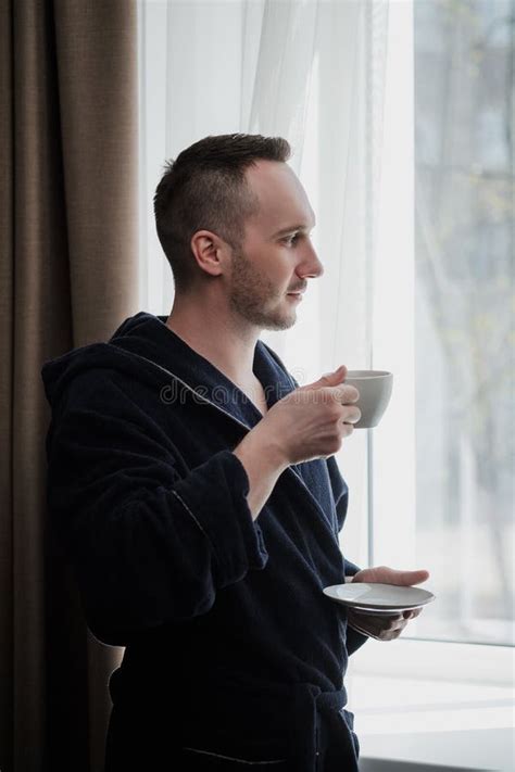 Young Brunette Man In A Blue Robe By The Window Drinks Coffee Or Tea