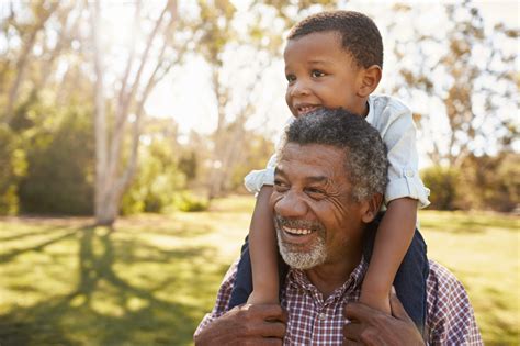 Grandfather Carries Grandson On Shoulders During Walk In Park Tell