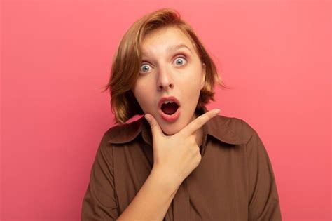 Surpris jeune fille blonde regardant mettre la main sur le menton isolé sur mur rose avec espace